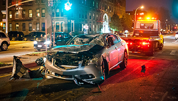 Severely damaged car at a nighttime crash scene with an emergency vehicle behind it.